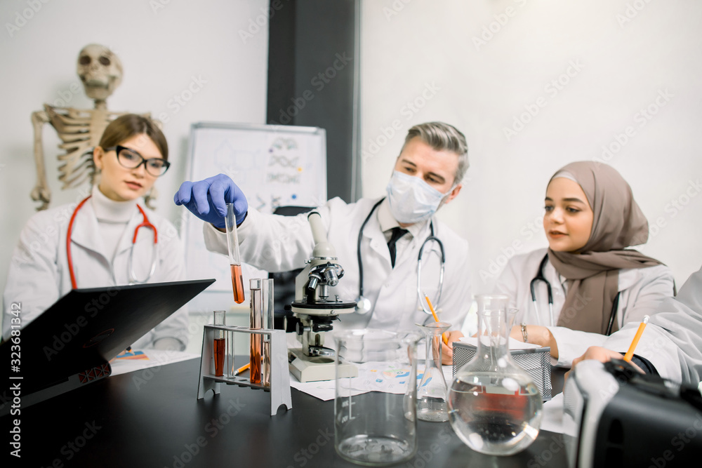 Medicine, genetics, immunology, microbiology concept. Multiethnic group of scientists, Caucasian man and woman, Muslim woman, working at the laboratory with microscope, tubes with liquids and laptop. Medicine, genetics, immunology, microbiology concept. Multiethnic group of scientists, Caucasian man and woman, Muslim woman, working at the laboratory with microscope, tubes with liquids and laptop.