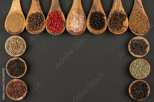 Seven cooking spoons made of olive wood and six small wooden bowls filled with various spices on a black background with copy space