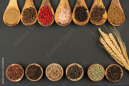 Seven cooking spoons made of olive wood in a row and six small wooden bowls in a row filled with various spices and grain ears on a black background with copy space