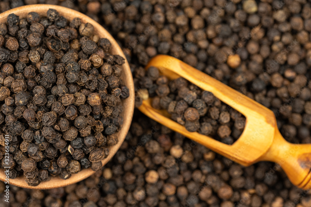 Spice background, Background made of many whole black peppercorns and a wooden bowl also filled with peppercorns and a spice shovel, close up, top view Spice background, Background made of many whole black peppercorns and a wooden bowl also filled with peppercorns and a spice shovel, close up, top view