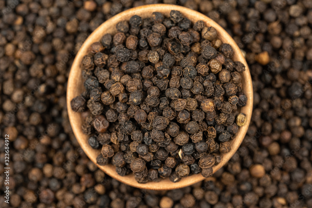 Spice background, Background made of many whole black peppercorns and a wooden bowl filled with peppercorns, close up, top view Spice background, Background made of many whole black peppercorns and a wooden bowl filled with peppercorns, close up, top view