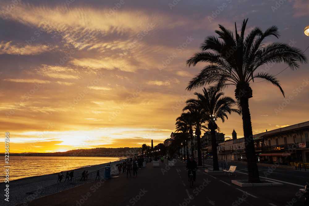 The sunset sun lights Promenade des Anglais - the main embankment of the city, one of the most beautiful in the world, and the city pebble beach. Brilliant sea beach sunset with palm trees Nice, Franc