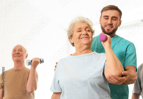 Care worker helping elderly woman to do exercise with dumbbell in hospital gym