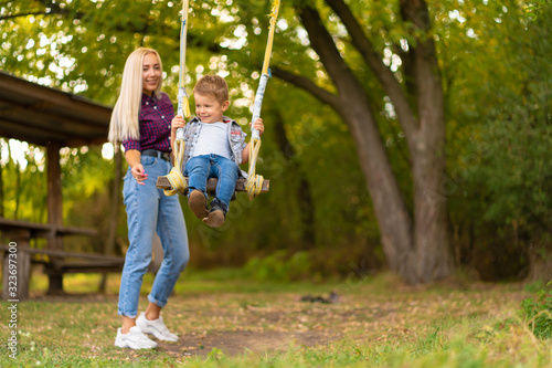 Young blonde mom shakes her little son on a swing in a green park. Happy childhood. © Kate