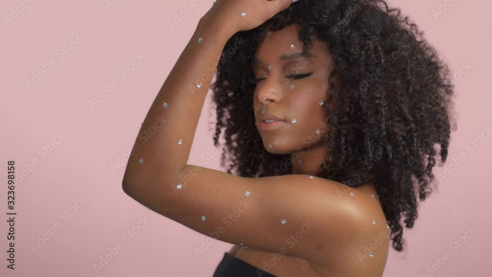 Mixed race black woman with curly hair covered by crystal makeup on pink background in studio Model moves her shoulder dancing Mixed race black woman with curly hair covered by crystal makeup on pink background in studio Model moves her shoulder dancing
