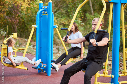 Beautiful family of three wearing sportswear spending time at the outdoor gym, focus on father working out on chest press machine, girls are training legs on background