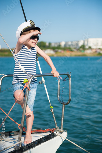 active little caucasian boy in a captain hat and sunglasses peers into distance standing on board of yacht during the summer sea cruise