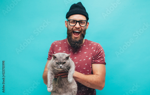 Handsome hipster guy with beard smiling happily, hugs his cute cat isolated over blue studio background. Animal lover. Friendship concept. photo