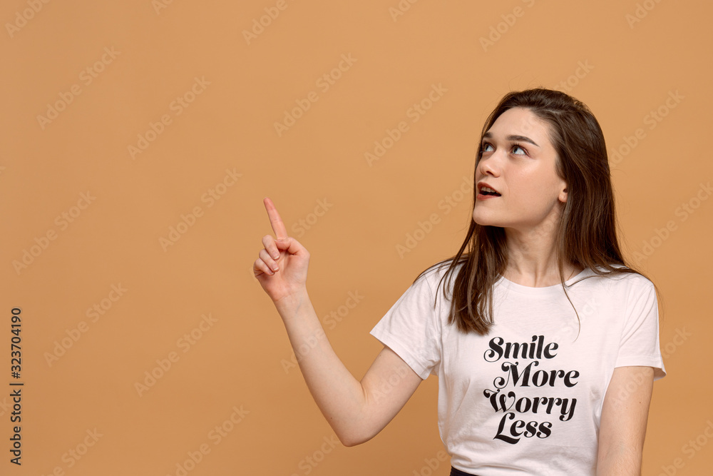 Pensive girl pointing with finger. Studio shot of emotional stylish woman in white t-shirt isolated over orange background with copyspace Pensive girl pointing with finger. Studio shot of emotional stylish woman in white t-shirt isolated over orange background with copyspace