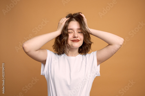 Lazy girl with shiny dark hair posing with hands up against orange background. Indoor portrait of curious young female model in white t-shirt