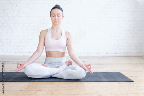 Healthy pacified young woman in tracksuit practices yoga in lotus position and mudra gesture on rug on floor on white brick wall. Healthy respiratory and nervous system concept. Advertising space