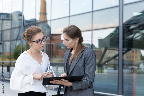 Two young and beautiful business women are standing against the background of an office building. They discuss the graphics that they saw on the tablet.
