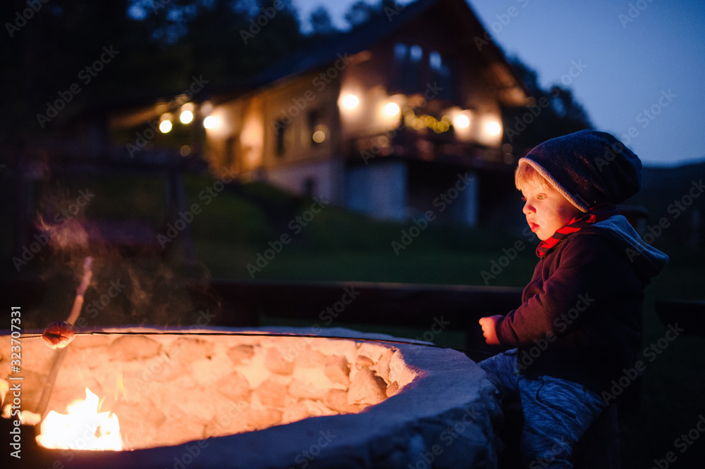 A toddler boy standing and playing by open fire outdoors in garden in summer.