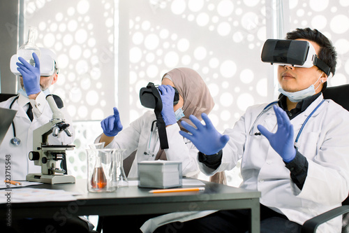 Team of scientists, Asian and Caucasian men, Muslim woman using virtual reality goggles for scientific research in laboratory, gesturing hands. Medicine, chemistry, microbiology, biotechnology concept
