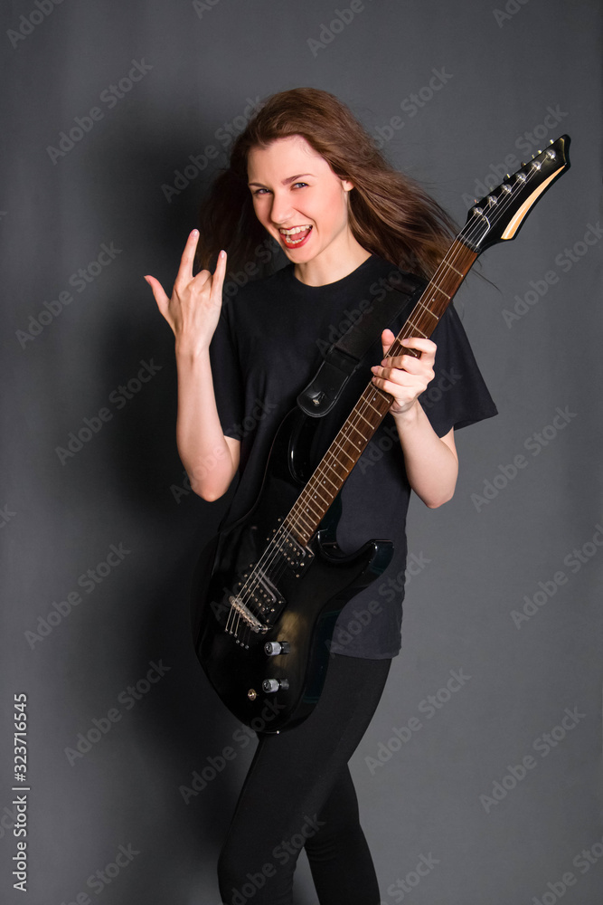 A beautiful, young rock girl in black clothes with an electric guitar in her hands shows a goat gesture. Studio photo on a gray background. Model with clean skin.