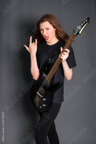 A beautiful, young rock girl in black clothes with an electric guitar in her hands shows a goat gesture. Studio photo on a gray background. Model with clean skin.