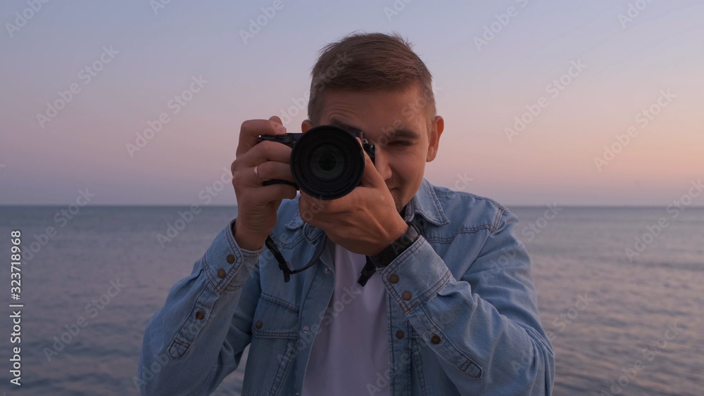 Obraz premium Portrait Of a male photographer in a denim shirt at sunset with a camera, the sea in the background.