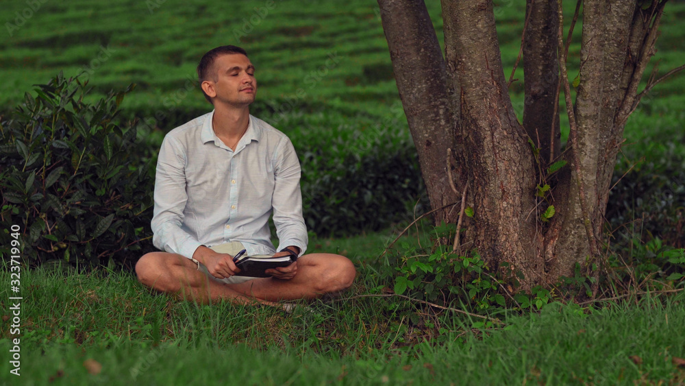 A young man with closed eyes sits on the grass under a tree with a notebook and enjoys nature. A young man with closed eyes sits on the grass under a tree with a notebook and enjoys nature.