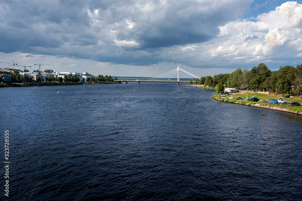 Obraz premium Republic of Finland, Rovaniemi: Kemijoki river next to the city center of the Finnish town with riverbank and modern white Jatkankynttila bridge (Lumberjack Candle Bridge) in background.
