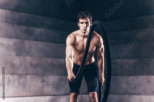 Handsome muscular man is doing battle rope exercise while working out in gym.