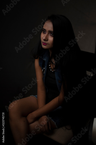 Fashion portrait of an young and attractive Indian Bengali brunette girl with tee shirt and blue western jacket sitting in front of a black studio background. Indian fashion portrait and lifestyle.