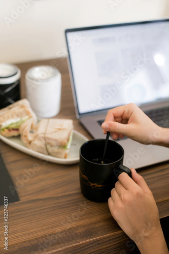 The girl sitting at a working wooden table with a laptop decided to have a snack and eat a hot tea sandwich in a black mug. Lunch break. The gadgets and technology that surround us.