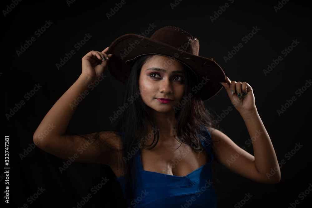 Fashion portrait of young and attractive Indian Bengali brunette girl with blue western dress with cowboy hat under spotlight in front of black studio background.Indian fashion portrait and lifestyle. Fashion portrait of young and attractive Indian Bengali brunette girl with blue western dress with cowboy hat under spotlight in front of black studio background.Indian fashion portrait and lifestyle.