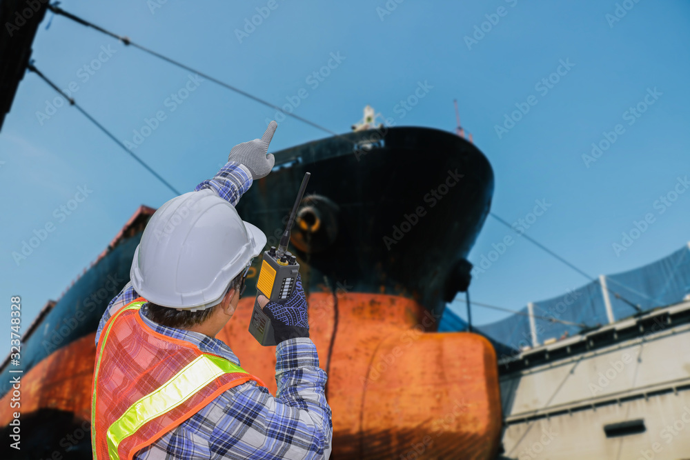 worker walkie talkie in hand operation control in Shipyard The bulk carrier general old cargo ship moored in dry dock yard of hull repairing and repainting, hand poiting target success concept. worker walkie talkie in hand operation control in Shipyard The bulk carrier general old cargo ship moored in dry dock yard of hull repairing and repainting, hand poiting target success concept.