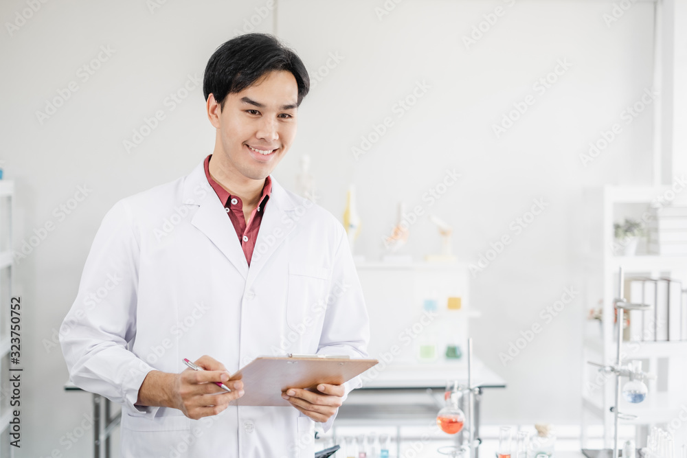 A male asian scientist smiling writing on a clipboard in white laboratory with test tube microscope and solutions. A male asian scientist smiling writing on a clipboard in white laboratory with test tube microscope and solutions.