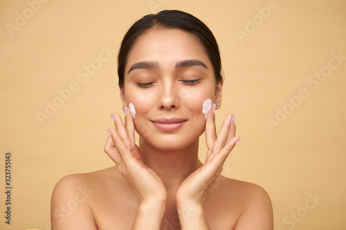  Young girl with closed eyes with clean skin puts cream on her face with hands on a yellow background