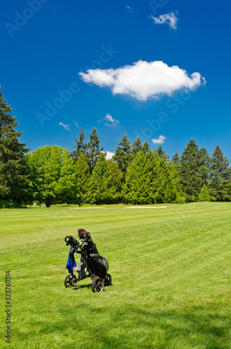 A golf bag on three wheel golf cart over a beautiful green with blue sky and white clouds.