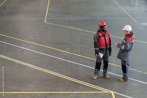 Horizontal high angle long shot of two young adult factory workers wearing uniform standing together chatting photo