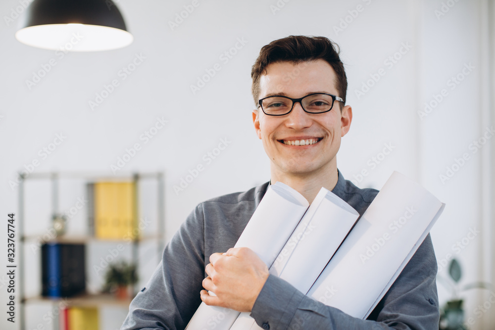 Attractive young man architect in glasses standing isolated over loft office background, carrying drawings, holding house scheme Attractive young man architect in glasses standing isolated over loft office background, carrying drawings, holding house scheme