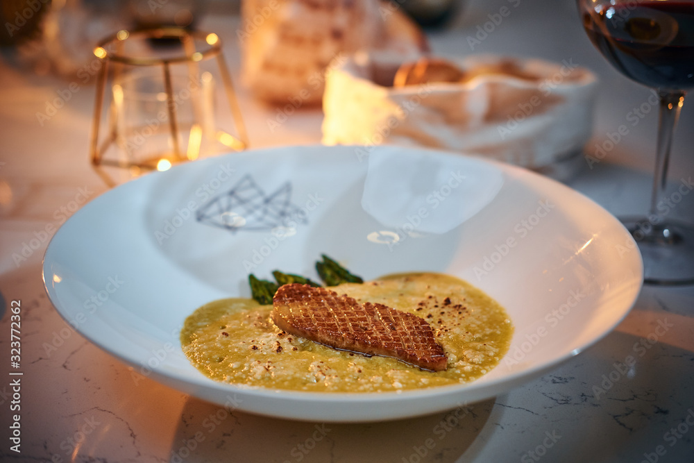 Excellent fillet of grilled and oven baked gourmet fish served with garnish including herbs and oil sauce, on a white porcelain dish next to golden candle, a glass of red wine and a full wicker basket Excellent fillet of grilled and oven baked gourmet fish served with garnish including herbs and oil sauce, on a white porcelain dish next to golden candle, a glass of red wine and a full wicker basket