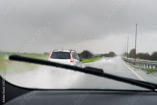 A car appearing clearly as a windscreen is wiped of rain by a wiper. Spray from cars can be seen on the wet road. Taken from inside the car looking out.