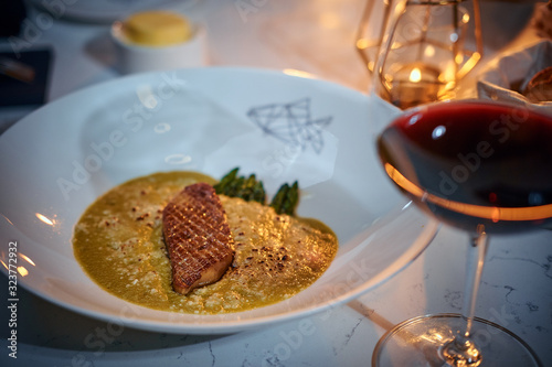Excellent fillet of grilled and oven baked gourmet fish served with garnish including herbs and oil sauce, on a white porcelain dish next to golden candle, a glass of red wine and a full wicker basket
