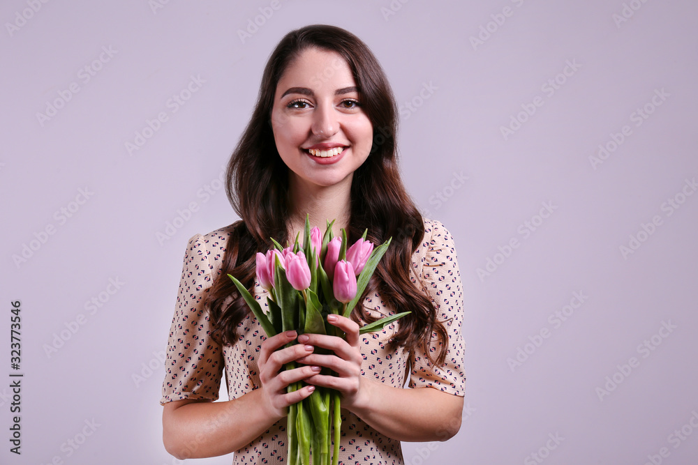Studio portrait of gorgeous young brunette woman with long wavy hair wearing slim cotton dress, holding bouquet of pink tulip flowers. Grey isolated background, copy space, close up. Studio portrait of gorgeous young brunette woman with long wavy hair wearing slim cotton dress, holding bouquet of pink tulip flowers. Grey isolated background, copy space, close up.