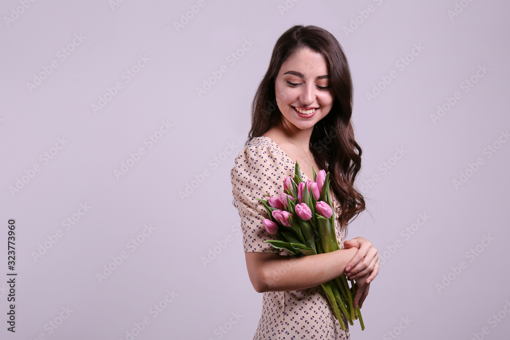 Studio portrait of gorgeous young brunette woman with long wavy hair wearing slim cotton dress, holding bouquet of pink tulip flowers. Grey isolated background, copy space, close up.