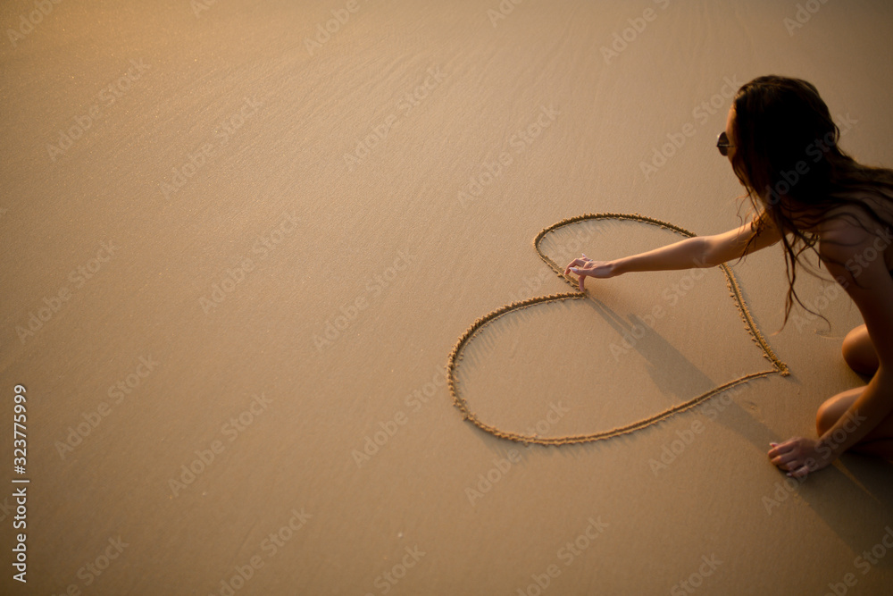 A beautiful slender girl in a swimsuit draws a heart on the sand with her finger near the sea at sunset in Thailand A beautiful slender girl in a swimsuit draws a heart on the sand with her finger near the sea at sunset in Thailand