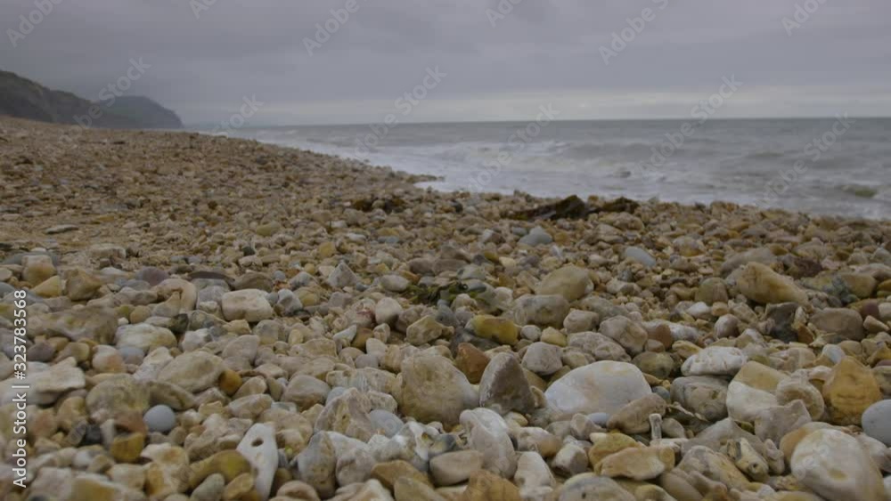 A medium, extreme-long shot of a rocky beach covered with stones pebbles of varied sizes and colours, with crashing waves and a mountain range in the background. A medium, extreme-long shot of a rocky beach covered with stones pebbles of varied sizes and colours, with crashing waves and a mountain range in the background.