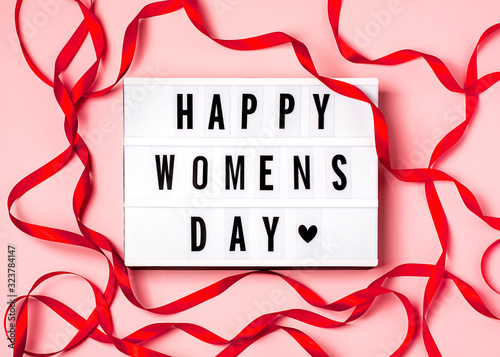 Happy women's day inscription on the lightbox, red ribbon on the pink background. 8 march concept. Top view, flat lay composition