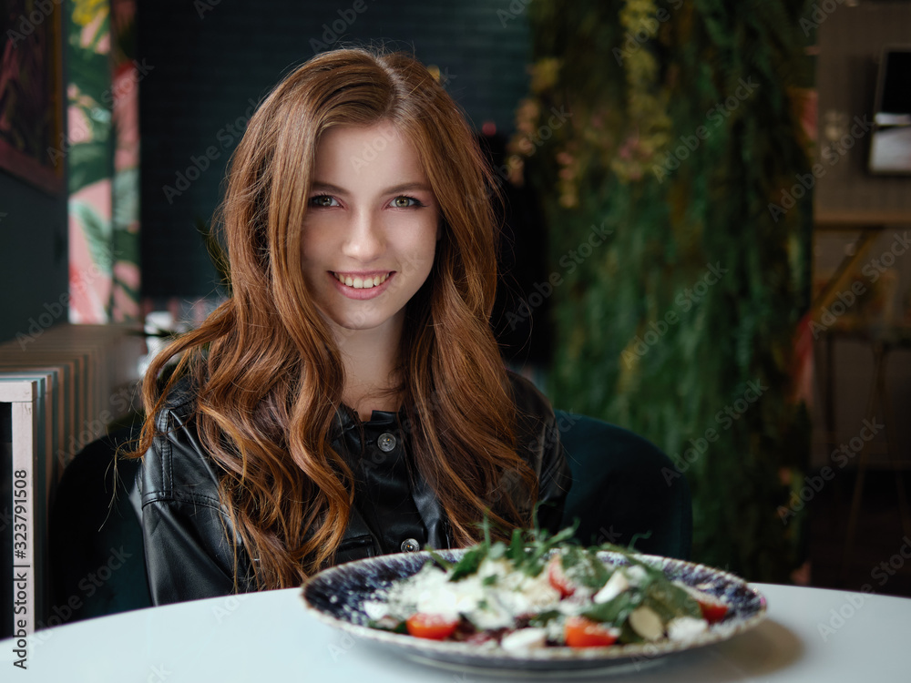 Close up shot of beautiful happy redhead woman in black leather shirt sitting in trendy cozy city cafe near big window with delicious salad on table