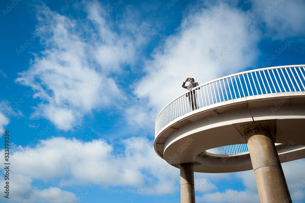 Distant businessman standing outdoors on curved walkway looking out in bright blue sky Distant businessman standing outdoors on curved walkway looking out in bright blue sky