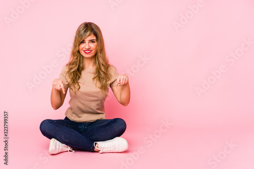 Young blonde caucasian woman sitting on a pink studio points down with fingers, positive feeling.