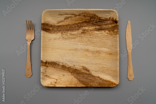 Textured disposable square plate made of bamboo with a wooden knife and fork on a neutral background.