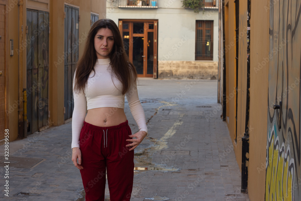 Portrait of caucasian young woman looking serious and pensive directly at camera, in the city, white top and red pants, dark air. Place for your text in copy space.