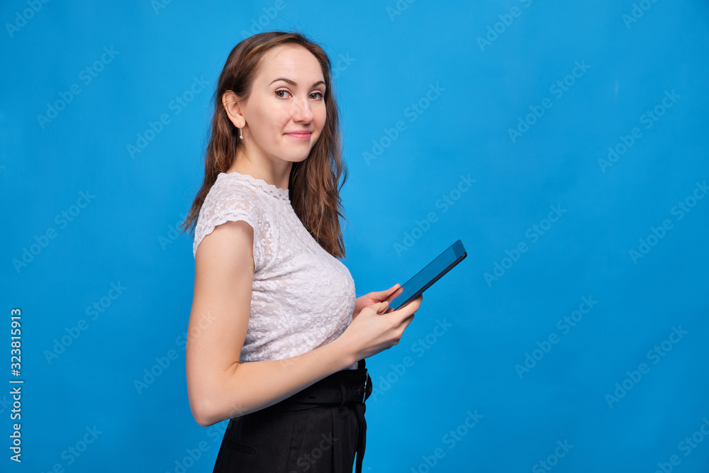 A brunette girl in casual clothes on a blue background and looks at the camera and holds a tablet. A brunette girl in casual clothes on a blue background and looks at the camera and holds a tablet.