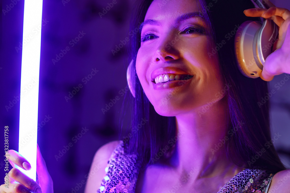 Toned portrait of young woman with headphones and neon lamp on dark color background