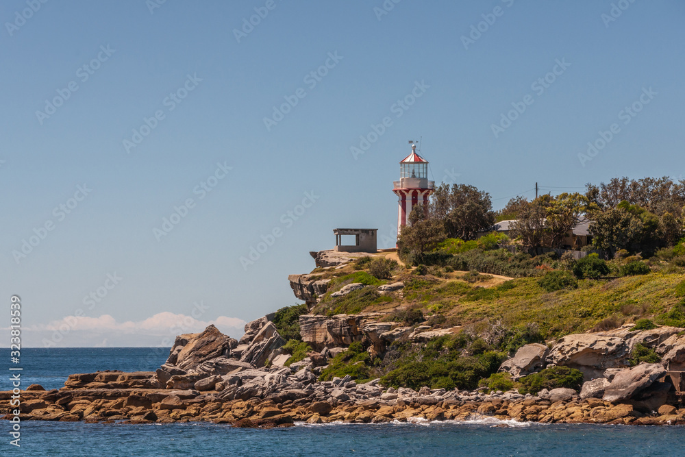 Sydney, Australia - December 11, 2009: Hornby lighthouse on brown and gray rocky cliff at entrance to bay under light blue sky. Deep blue sea water.