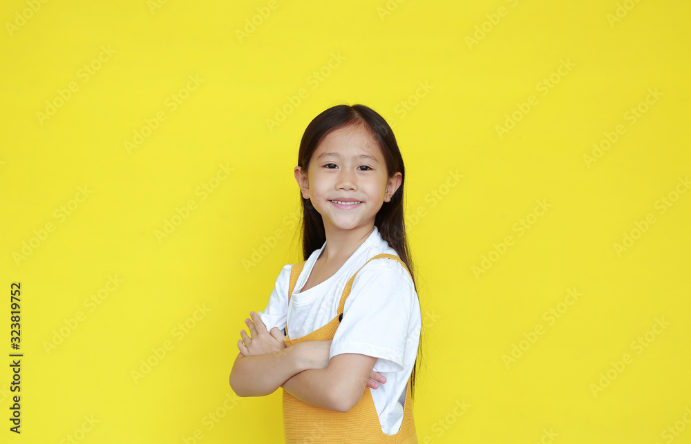 Smart kid girl crossed arms with face smiling looking at camera isolated on yellow background. Little asian child in dungarees Smart kid girl crossed arms with face smiling looking at camera isolated on yellow background. Little asian child in dungarees
